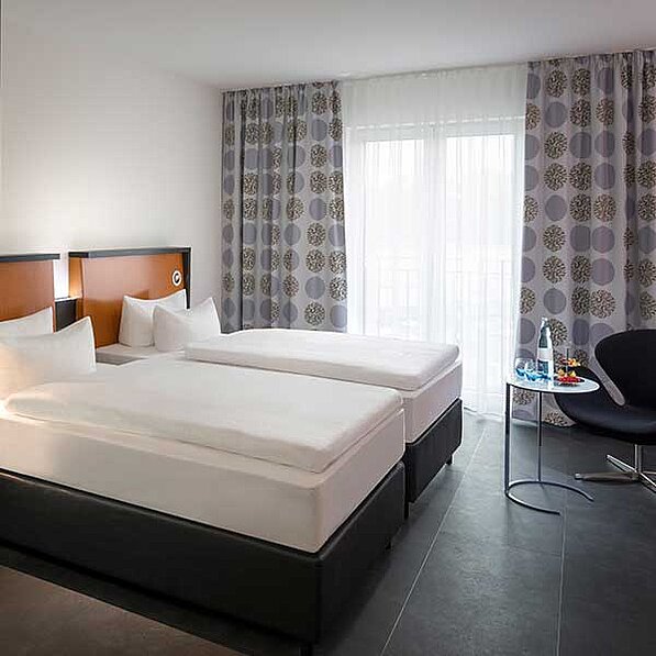 Im Bild ist ein modernes Hotelzimmer mit zwei Einzelbetten zu sehen, die zusammengefügt sind und so ein Doppelbett bilden. Die Betten haben weiße Bettwäsche und stehen auf schwarzen, niedrigen Bettrahmen. An den Wänden befinden sich orangefarbene Kopfteile. Die Fenster sind mit hellen Vorhängen und grauen Vorhängen mit einem Muster aus stilisierten Kreisen versehen. Ein kleiner, runder Beistelltisch steht zwischen den Betten und trägt ein Glas Wasser, ein paar Gläser und eine kleine Schale mit möglicherweise Obst. Ein schwarzer, moderner Stuhl steht rechts neben dem Tisch. Der Boden ist dunkelgrau gefliest. Das Gesamtbild wirkt sauber, modern und ruhig. Die Atmosphäre ist entspannend und einladend, typisch für ein Hotelzimmer.
