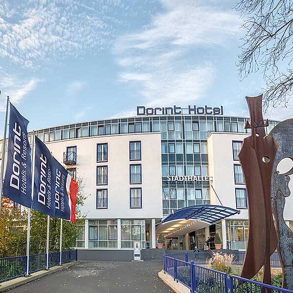 Das Bild zeigt ein modernes, weißes Hotelgebäude mit großen Fenstern. Über dem Eingang prangt das Schild „Dorint Hotel“. Vor dem Hotel stehen mehrere Fahnenmasten mit Dorint-Fahnen, die das Hotel und seine Zugehörigkeit zu einer Hotelkette kennzeichnen. Der Eingangsbereich ist mit einem Vordach versehen und wirkt einladend. Rechts neben dem Eingang steht eine abstrakte, rostbraune Skulptur, die Aufmerksamkeit auf sich zieht. Im Hintergrund ist ein Teil der Stadthalle sichtbar, wie ein Schild oberhalb des Hoteleingangs zeigt. Der Himmel ist leicht bewölkt, das Licht erscheint weich und diffus. Die allgemeine Atmosphäre ist ruhig und gepflegt, das Bild vermittelt einen Eindruck von modernem Komfort und professionellem Service. Im Vordergrund ist ein Teil eines Zaunes mit einem Gehweg zu sehen. Einige Personen sind im Eingangsbereich des Hotels zu erkennen, jedoch nicht im Detail. Die Bäume im Hintergrund sind kahl, was auf die Jahreszeit (Herbst oder Winter) hindeuten könnte. Das Bild wirkt insgesamt ruhig und professionell, typisch für die Präsentation eines Hotels.