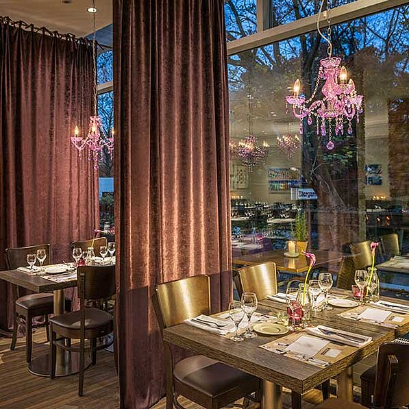 Im Detail zeigt das Bild zwei Essgruppen, die jeweils mit mehreren Stühlen und einem Tisch aus dunklem Holz ausgestattet sind. Auf den Tischen stehen Gedecke, Gläser und Vasen mit Blumen. Über den Tischen hängen zwei Kristallleuchter, die einen Hauch von Luxus verleihen. Die Fenster zeigen eine Aussicht auf einen grünen Außenbereich mit Bäumen und Büschen, und im Hintergrund sind weitere Tische und Stühle zu sehen, die auf andere Essbereiche im Restaurant hindeuten. Die allgemeine Stimmung des Bildes ist warm, einladend und entspannend mit einer Mischung aus Eleganz und Gemütlichkeit. Die Beleuchtung im Raum scheint gedämpft, was die Stimmung des Bildes ebenfalls verstärkt.